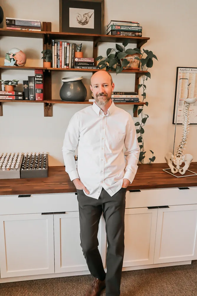 Dr. Andrew Rostenberg, DC, DIBAK standing in his Boise treatment room in front of a wooden bookshelf of medical reference books, with an Applied Kinesiology spine model and charts visible