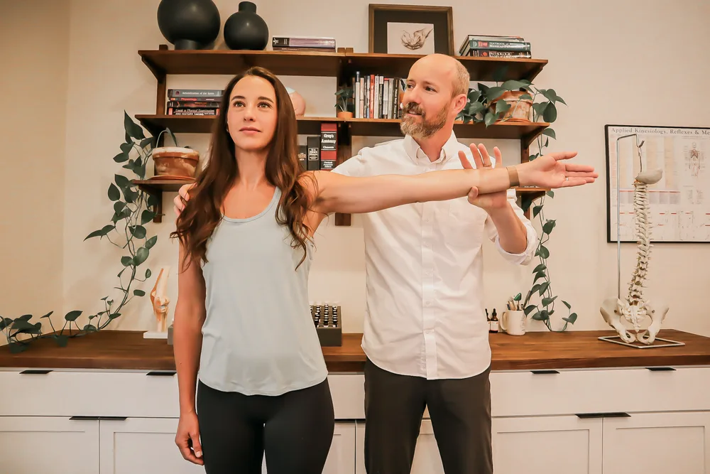 Dr. Andrew Rostenberg performing applied kinesiology muscle testing — arms-outstretched bilateral test — on a patient at Red Mountain Natural Medicine in Boise, Idaho, with anatomy books, spine model, and AK reference chart visible behind
