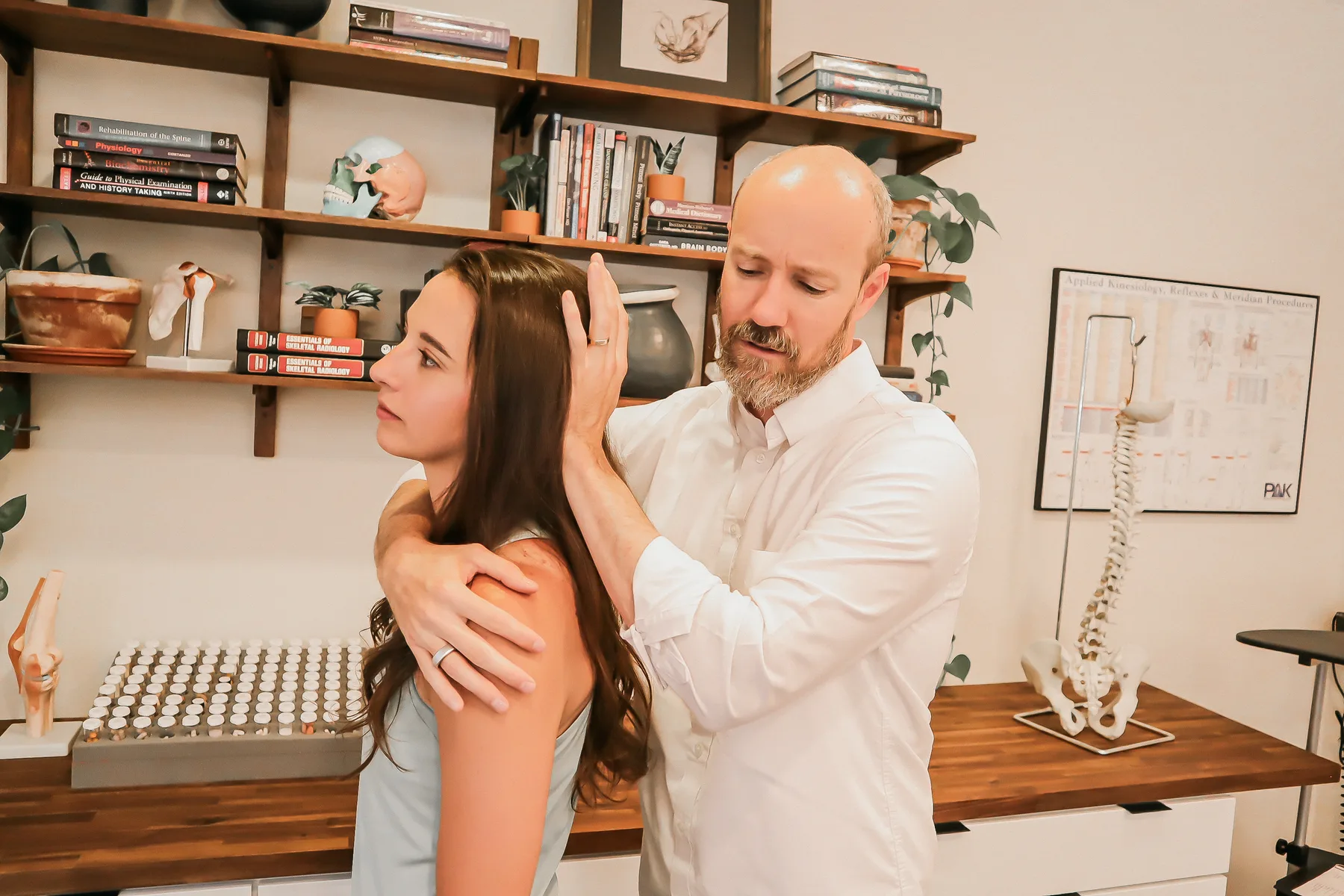 Dr. Andrew Rostenberg, DC, DIBAK performing an applied kinesiology cervical and upper-cervical evaluation on a patient at Red Mountain Natural Medicine in Boise, Idaho — anatomy books, spine model, and AK reference chart visible behind