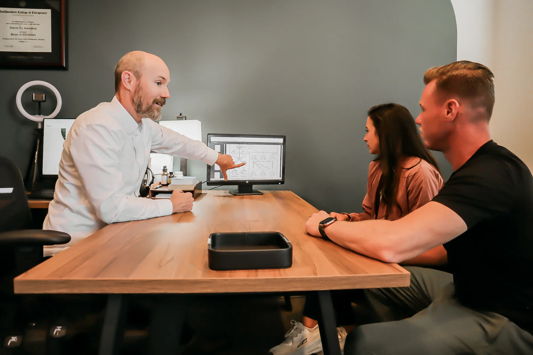 Dr. Andrew Rostenberg walking patients through findings on a monitor at Red Mountain Natural Medicine in Boise, Idaho — framed credentials and chiropractic diplomas on the wall behind