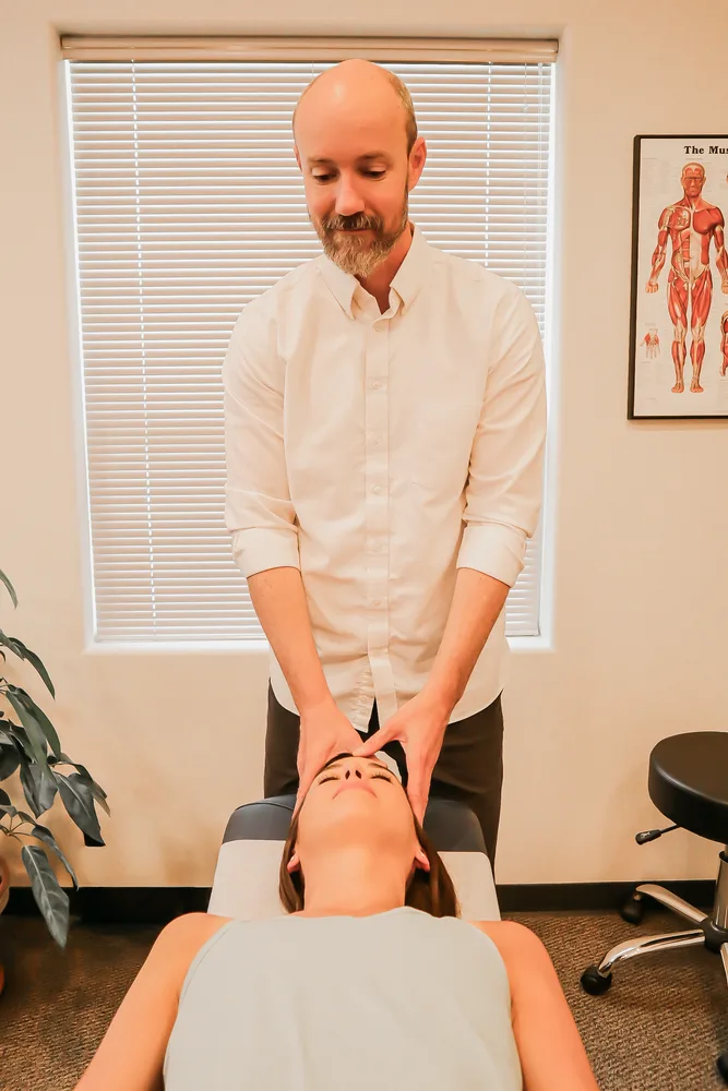 Dr. Andrew Rostenberg performing a gentle cranial hold on a patient on the adjustment table at Red Mountain Natural Medicine in Boise, Idaho — the muscular anatomy chart visible on the wall behind
