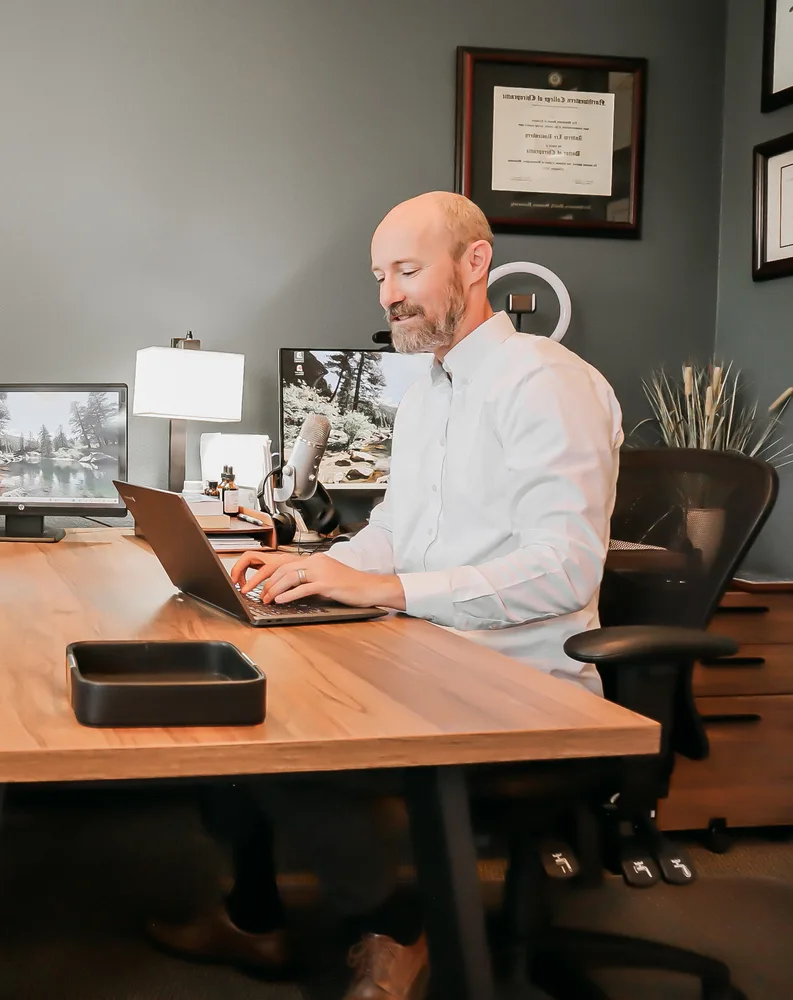 Dr. Andrew Rostenberg seeing a patient over telehealth from his desk at Red Mountain Natural Medicine in Boise, Idaho — laptop, professional microphone, ring light, and his Doctor of Chiropractic diploma on the wall behind him